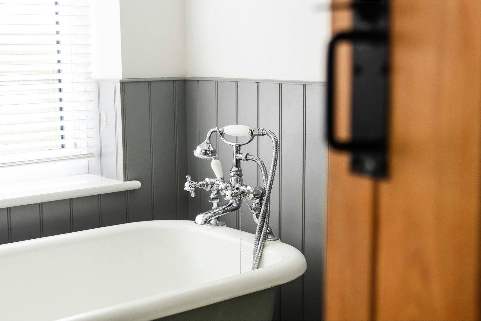 A modern bathroom with grey panelling and a roll top bath with an oak door