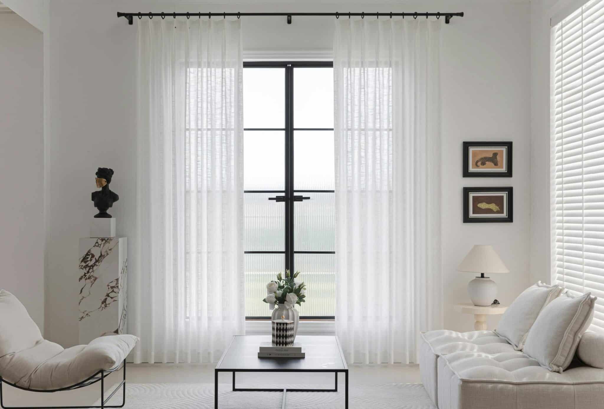 A white and minimalist living room with sheer curtains on one window and blinds on another
