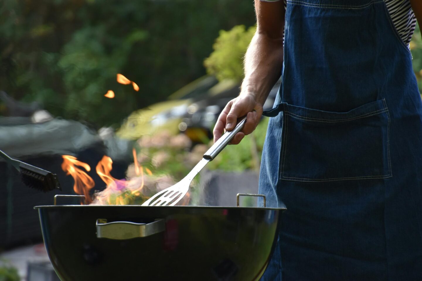 A person wearing an apron and using a spatula to turn food on a charcoal barbeque